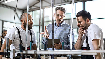 Zwei junge Fabrikmitarbeiter sehen sich in Fertigungshalle mit ihrem älteren Kollegen digitale Auswertungen einer Maschine auf Tablet an und ziehen daraus wertvolle Schlüsse für ihre Produktion
