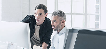 LANCOM Anregungen zur Informationssicherheit für mittelständische Unternehmen Junger IT-Mitarbeiter mit Zopf berät Kollegen mittleren Alters mit grauen Haaren und Bart zu Sicherheitseinstellungen am PC