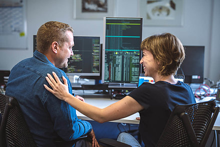 LANCOM WLAN-Produkte – für ein WLAN, bei dem alles passt Arbeitssituation mit Blick von hinten über die Schultern zweier Mitarbeitenden: Junge Frau in T-Shirt mit kurzen, karamellfarbenen Haaren klopft ihrem blonden Kollegen in Hemd anerkennend auf die Schulter; im Hintergrund sind mehrere PC-Bildschirme mit Programmiersprache darauf auf einem Schreibtisch zu sehen